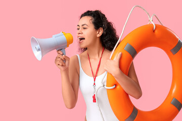 Beautiful young African-American female lifeguard with ring buoy and megaphone on pink background