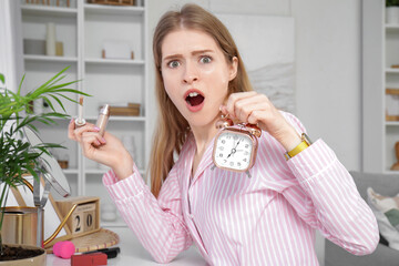 Shocked young woman with alarm clock applying makeup at home