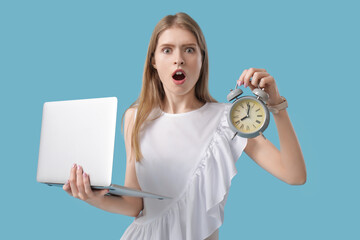 Shocked young woman with alarm clock and laptop on blue background