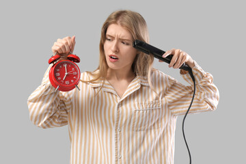 Hurrying young woman in pajamas with hair straightener checking time on alarm clock against grey...