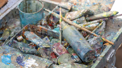 An outdoor artist's workspace is captured, showcasing a well-used metal box with a wooden panel, paintbrushes, and cans of paint, all covered in vibrant splatters, reflecting the creative process.