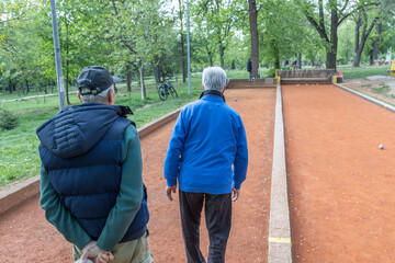 In a serene park setting, two older men engage in a game of boules. One man, dressed in a blue...