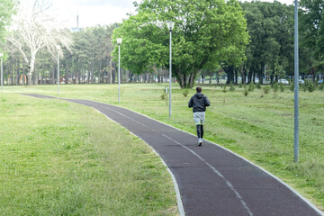  A solitary figure is captured in motion, jogging along a paved path in an urban park. The park is...