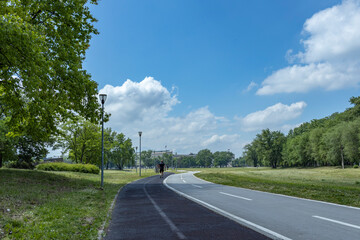 A serene bike path lined with lush trees and grass, under a blue sky with scattered clouds, where a...