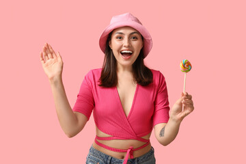 Surprised young woman with sweet lollipop on pink background