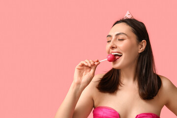 Beautiful young woman in party hat with sweet lollipop on pink background