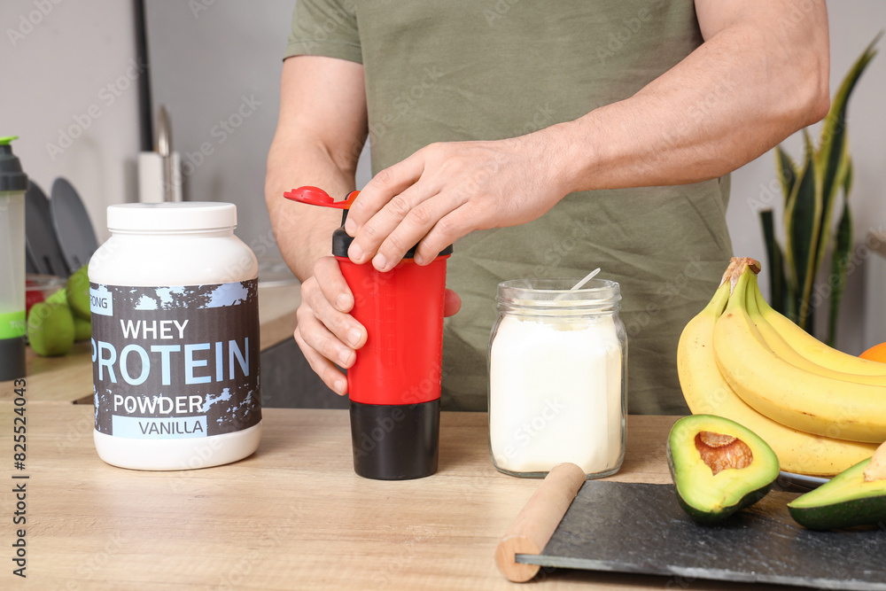 Sticker Sporty muscular man making protein shake at table in kitchen, closeup