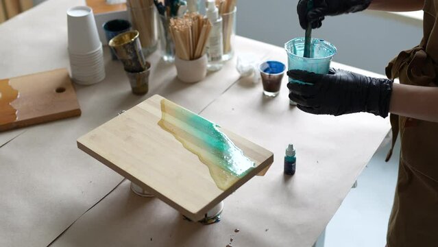 Creative craftswoman adds colorant blue color in fluid epoxy, mix slowly. Process making handmade stylish craft closeup using modern materials for design, acrylic, polymers, natural wood and stone.
