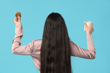 Beautiful young woman holding tasty cookie with chocolate chips and glass of milk on blue...