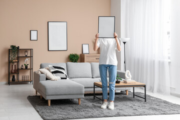 Young woman holding picture near grey sofa in living room