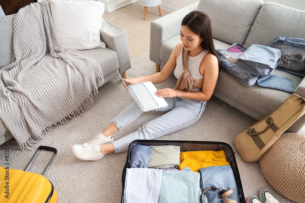 Wall mural Female Asian traveler using laptop while packing suitcase at home