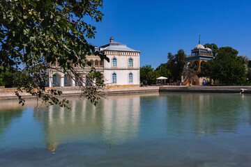 Scenic view of Emir Summer Palace in Bukhara, reflected in a tranquil pond, framed by lush greenery and a clear blue sky. Bukhara, Uzbekistan
