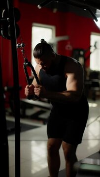 Muscular man with a beard and shaved head, wearing a black tank top and shorts, performing tricep pushdowns in a gym with red walls and exercise equipment