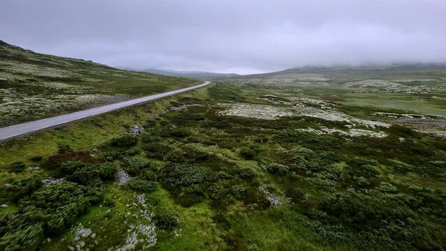 Drone flying parallel to a empty road in foggy tundra mountain landscape overgrown with green mossy vegetation in Rondane National Park, Innlandet, Norway.