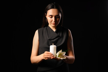Young woman with funeral lily flower and burning candle on black background