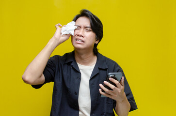 asian man crying over the phone after receiving bad news. Young asian man wiping tears with tissue...