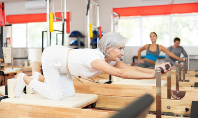 Senior woman visitor of fitness club is engaged in Pilates simulator. Improvement of well-being, physical condition, development of attractive body and figure during lessons at reformer.