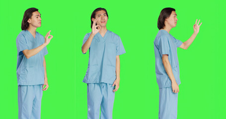 Obraz premium Medical specialist presents okay gesture over greenscreen backdrop, showing approval and like symbol. Healthcare worker in uniform doing agreement sign standing in studio.