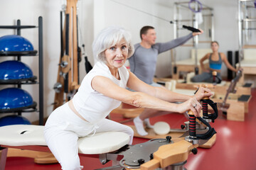 Elderly woman in sportswear stretches her arms and shoulders using pilates machine..