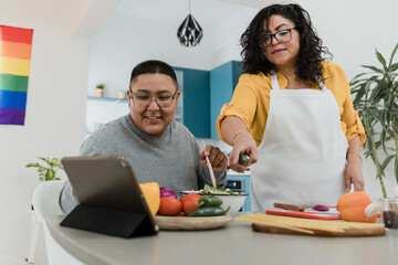 Latin lesbian couple cooking dinner at home in Mexico, Hispanic homosexual people from lgbt community with rainbow flag in Latin America