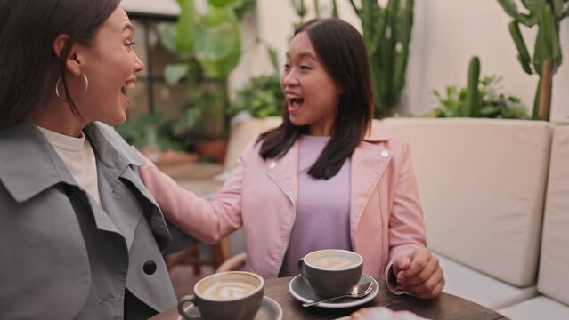 Asian woman affectionately kisses her friend on the cheek as they greet at a welcoming, cozy cafe, expressing happiness and friendship over coffee.