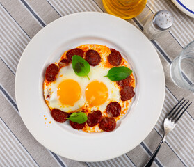 bright breakfast of sunny side up eggs with slices of chorizo sausage decorated with fresh green basil leaves is served on a white plate
