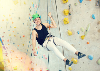 Middle-aged man in safety helmet and with safety rope hangs near climbing wall in gym. active hobby...