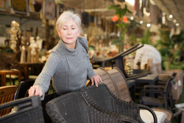 Interested elderly woman visiting furnishing store looking for practical plastic wicker chairs for...