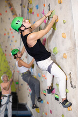 young positive man, insured by trainer, moving up artificial climbing wall indoors. Fun active...