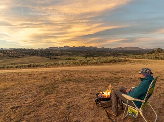 Mature caucasian man sitting in front of a fire watching the sunset over distance mountains, Lander Cutoff, Wyoming © Ron