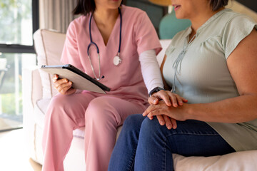 At home, Asian nurse in pink scrubs holding tablet, listening