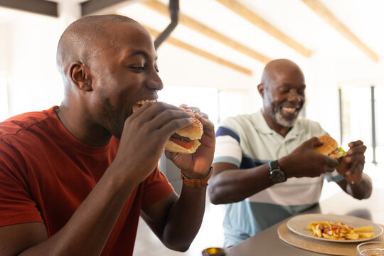African American senior father and adult son eating burgers at home