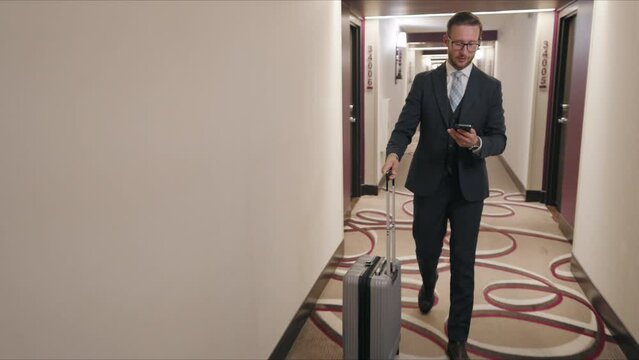 Businessman checks his smartphone while walking out from his room with a suitcase in a hotel corridor, illustrating a life always connected, even during travel. Slow motion. 
