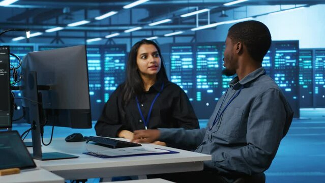 Multiracial team of IT engineers in server room brainstorming ways to fix equipment storing datasets. Indian and african american coworkers discussing how to mend racks doing computational operations
