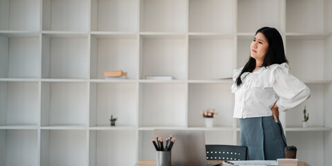 A woman is standing in front of a white wall with a laptop on a desk. She is wearing a white shirt...