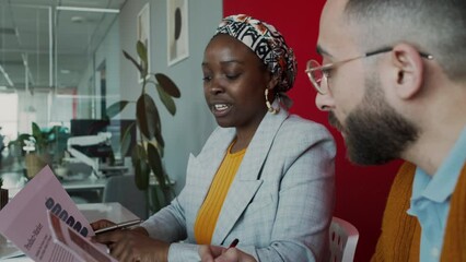Medium close-up of stylish young black woman in jacket and ethnic turban and Middle Eastern man in glasses discussing marketing research presentation slides in corporate meeting room