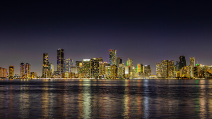 Obraz premium Night view of a Miami city skyline reflected on a calm body of water, showcasing the illuminated buildings against a dark sky. Ideal for themes of urban nightlife and modern cityscapes.