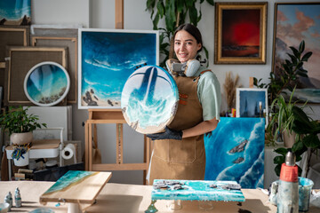 Satisfied girl artist holds finished epoxy resin painting, look to camera show off work in small...