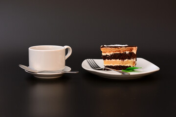 A piece of fresh chocolate biscuit cake on a plate decorated with mint leaves, a fork and a cup of hot black coffee on a black background.