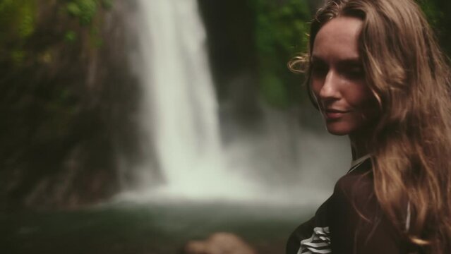 Woman standing in front of powerful Bali waterfall, surrounded by lush greenery. The water cascades down creating a dramatic backdrop for the serene moment. Air Terjun Munduk, Melanting. Slow motion