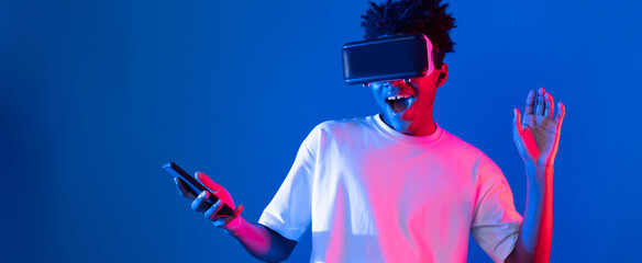 Young African American player wearing VR with using smartphone playing game hologram on blue pink neon light wall connecting digital futuristic technology virtual reality metaverse world. Contrivance.