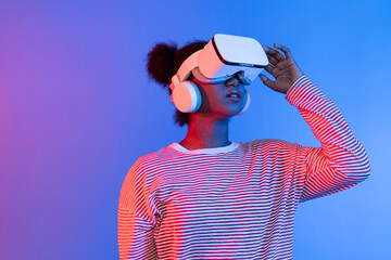 Smiling African woman wearing VR smelling good through metaverse flowers filed world and butterfly insect bug hologram vibrant neon pink blue lighting background technology 3D cyberspace. Contrivance.