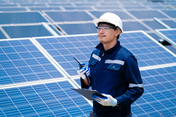 A man wearing a blue jacket and white hat is standing in front of a solar panel. He is holding a...