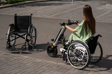 Woman in wheelchair with assistive device for manual control next to classic wheelchair. Electric...
