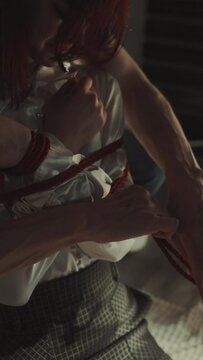 Shibari session in progress, featuring red rope binding woman's torso and arms, while another person tightens the rope. Intricacy and intimacy of traditional Japanese bondage art