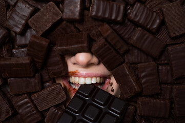 The face of a caucasian woman surrounded by chocolates. The girl eats a bar of chocolate.