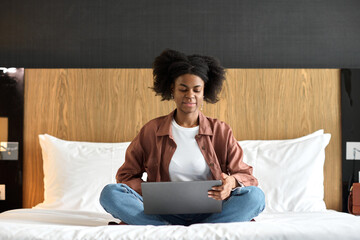 Portrait of young African American woman using laptop in hotel room while sitting on bed with legs...