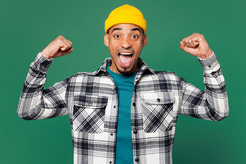 Young strong man of African American ethnicity he wear shirt blue t-shirt yellow hat showing biceps muscles on hand demonstrating strength power isolated on plain green background. Lifestyle concept.