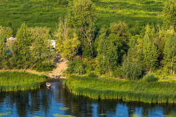 Quiet backwater overgrown with reeds, small fishing boat on the Volga River near the city of Samara, Russia. Blue water, sandy shore, tree, forest, bush. Quiet summer morning with light sky.