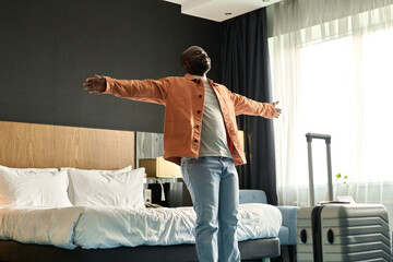 Portrait of joyful Black man falling onto bed with arms spread arriving in luxury hotel copy space